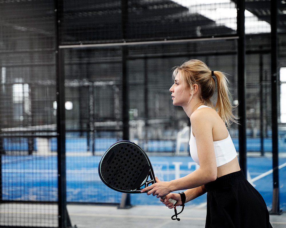 Joueuse concentrée tenant sa raquette sur un terrain de padel couvert, entourée de plusieurs pistes au sein d’un complexe sportif.